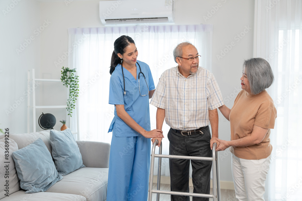 Fototapeta premium Asian young woman caregiver doctor during home visit helping elderly man patient to walk using walker frame while older woman his wife sitting on sofa encourage to him