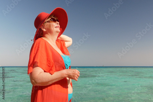 Happy middle aged woman wearing in swimsuit, big red sun hat and glasses staying on sea background in sunny summer day, holiday and travel concept photo