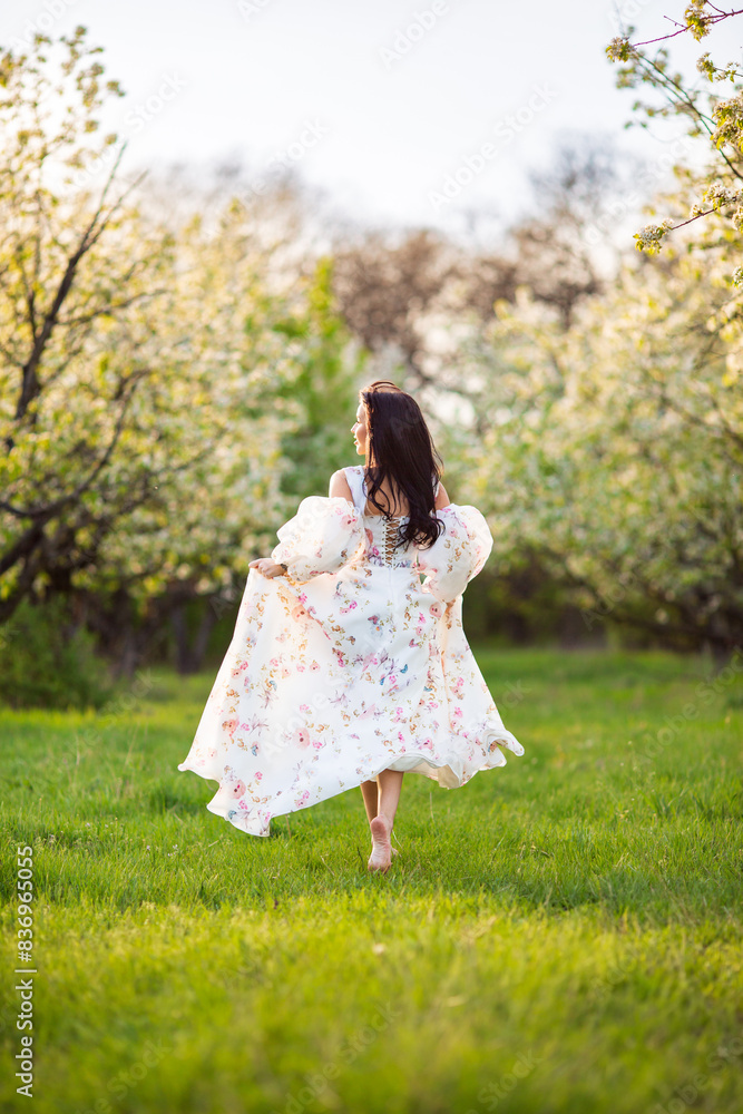 Full-length portrait of a beautiful brunette caucasian woman running in blossoming apple garden in white floral dress on a fresh green grass shot from her back with her hair flying Full-length portrait of a beautiful brunette caucasian woman running in blossoming apple garden in white floral dress on a fresh green grass shot from her back with her hair flying