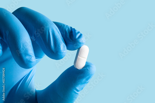 Blue-gloved hand holding a white pill against a light blue background.