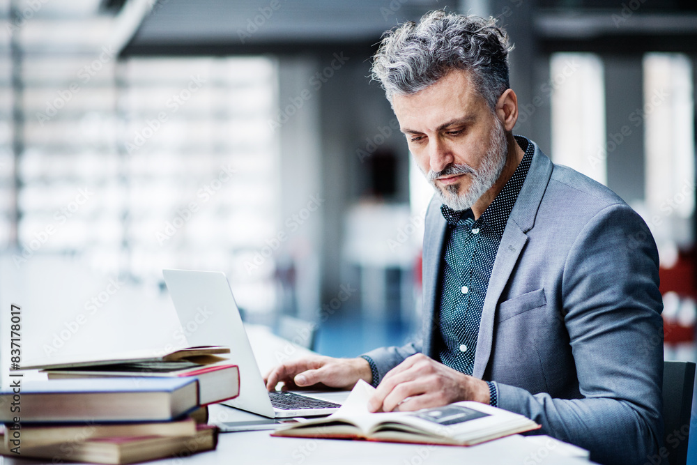 Handsome businessman working on laptop, searching for informations in book. Traditional information source in modern office space. Handsome businessman working on laptop, searching for informations in book. Traditional information source in modern office space.