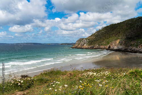 Plage enserrée par les falaises, végétation printanière en premier plan, mer d'Iroise turquoise, ciel bleu parsemé de nuages blancs, journée idyllique sur la presqu'île.