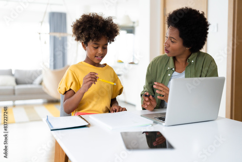 Supportive mother helping her daughter to finish her homework. She is explaining something while the girl is writing in a notebook. They are also using laptop for school work.