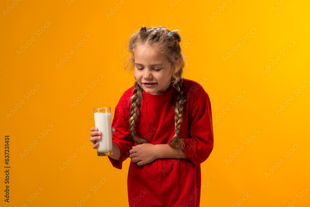 Kid girl holding glass of milk and feeling abdominal pain on yellow background. Lactose intolerance concept