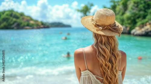Back view of attractive woman in a bikini and straw hat on idyllic tropical beach facing the ocean. Travel and summer vacation concept