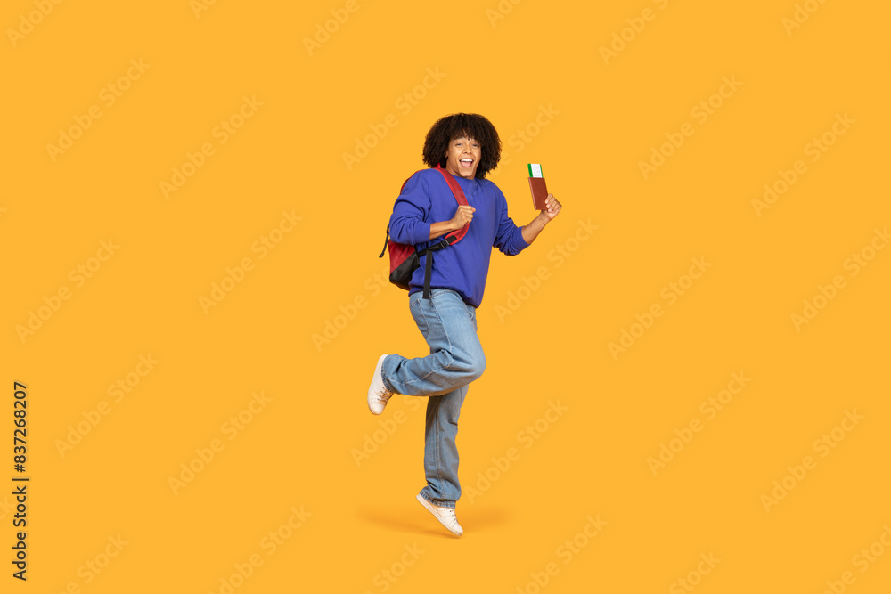 Fototapeta premium A cheerful guy wearing a blue sweater and carrying a red backpack jumps in excitement while holding a passport and ticket. The vibrant orange background adds energy and optimism to the scene.