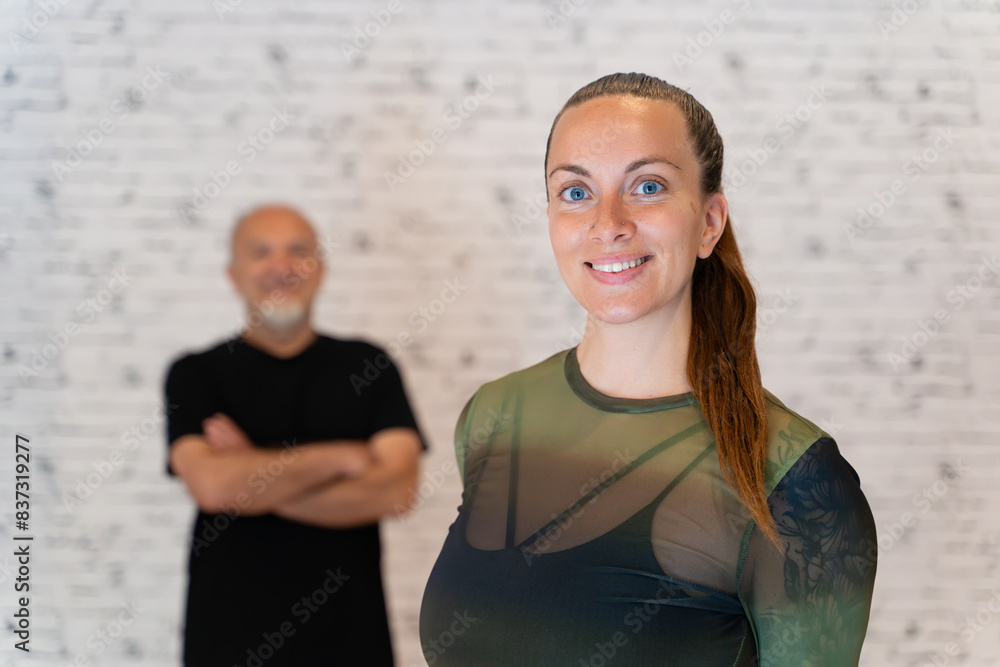 Smiling young woman in a sheer green top in the foreground with an elderly man in a black shirt in the background, both against a white brick wall. 
