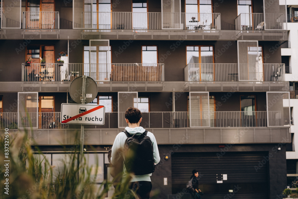 Young man with backpack walking in courtyard of modern European residential building, back view Young man with backpack walking in courtyard of modern European residential building, back view