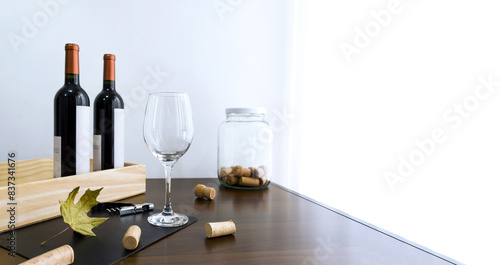 Copa de cristal vacía con botellas de vino tinto, sacacorchos y corchos dispersos sobre la mesa.  photo