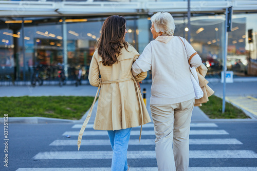 A young woman guides her older mother across a zebra crossing, both carrying grocery bags after shopping, illustrating family support in daily activitie