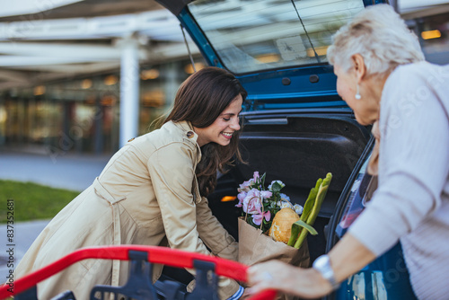 Foto A Caucasian adult daughter is cheerfully loading groceries into the car trunk, assisting her elderly mother outside a shopping center