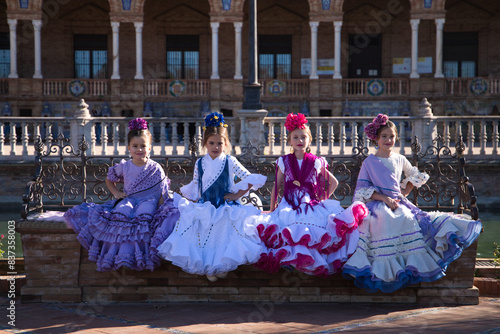 Four little girls dancing flamenco dressed in typical flamenco dress sitting on a bench in a famous square in seville, spain. In the background the arches and columns of the famous square.