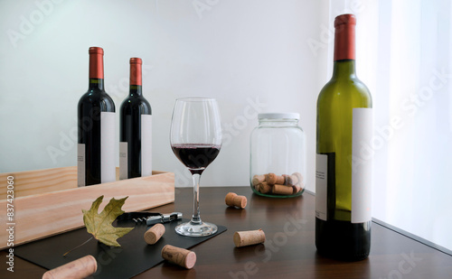 Copa de vino tinto servida junto a botellas de vino, sacacorchos y corchos dispersos sobre mesa de madera.    photo