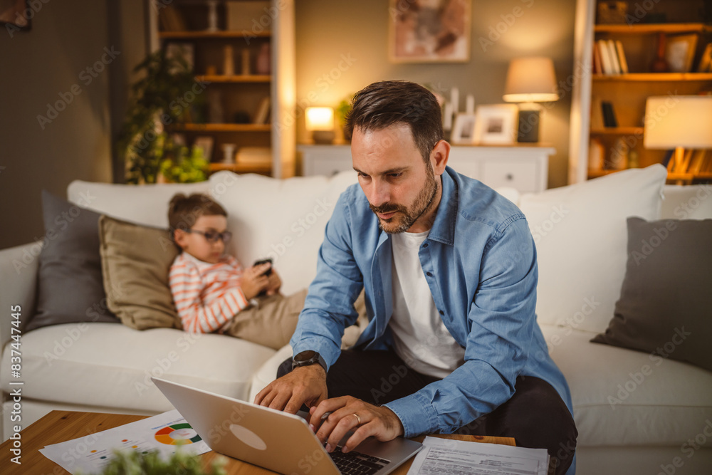 Fototapeta premium Father type on the lap top while son lie down on the sofa