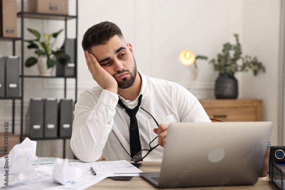 Overwhelmed man sitting at table with laptop and documents in office Overwhelmed man sitting at table with laptop and documents in office