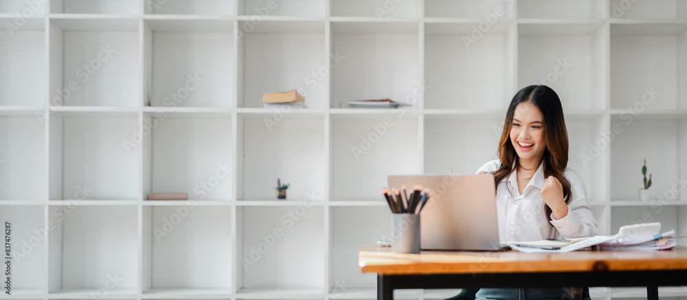 Young Professional Woman Working on Laptop in Modern Office with Minimalist Bookshelf Background, Smiling and Celebrating Success, Business and Productivity Concept Young Professional Woman Working on Laptop in Modern Office with Minimalist Bookshelf Background, Smiling and Celebrating Success, Business and Productivity Concept