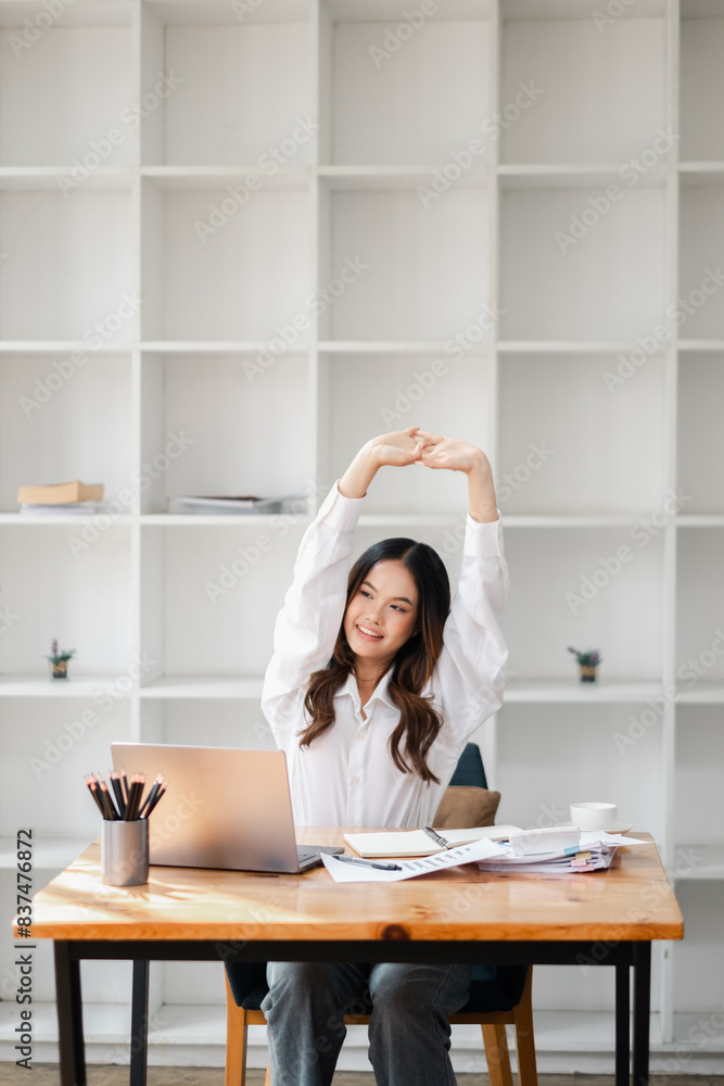 Young Professional Woman Stretching at Desk in Modern Office with Laptop and Documents, Relaxing During Work Break, Bright and Minimalistic Workspace with Shelves in Background Young Professional Woman Stretching at Desk in Modern Office with Laptop and Documents, Relaxing During Work Break, Bright and Minimalistic Workspace with Shelves in Background