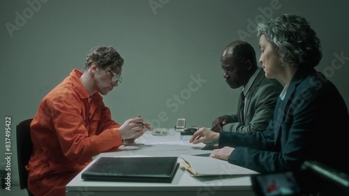 Medium side shot of diverse detectives interrogating murder suspect, filming on video camera. Caucasian female agent giving detainee in orange prison robe confession letter, man signing it photo