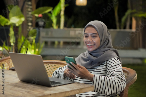 Southeast Asian hijab woman using her smartphone and laptop computer at outdoor cafe at night