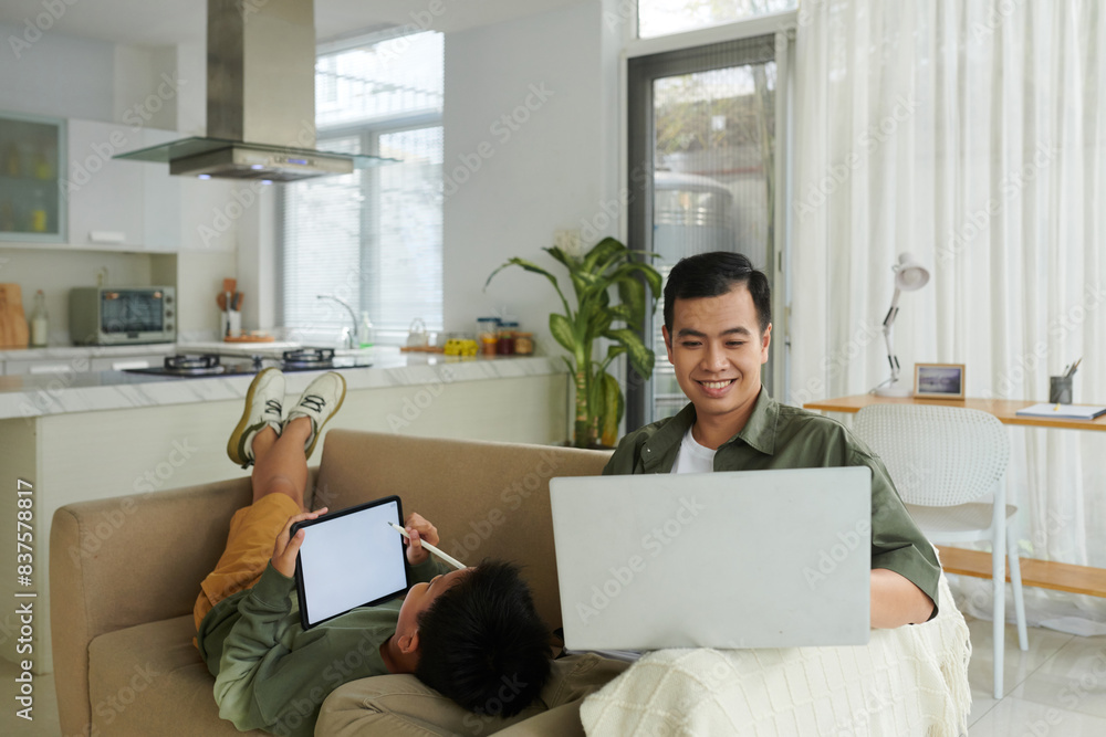 Smiling man working on laptop from home when son drawing on tablet screen Smiling man working on laptop from home when son drawing on tablet screen