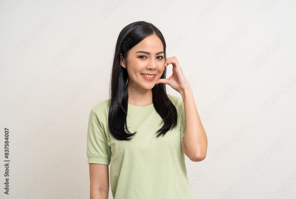 Pretty Young beautiful Asian woman standing and smiling showing mini heart sign on cheek with isolated white background. Lovely Latin female wearing green shirt feeling surprised and happy. Pretty Young beautiful Asian woman standing and smiling showing mini heart sign on cheek with isolated white background. Lovely Latin female wearing green shirt feeling surprised and happy.