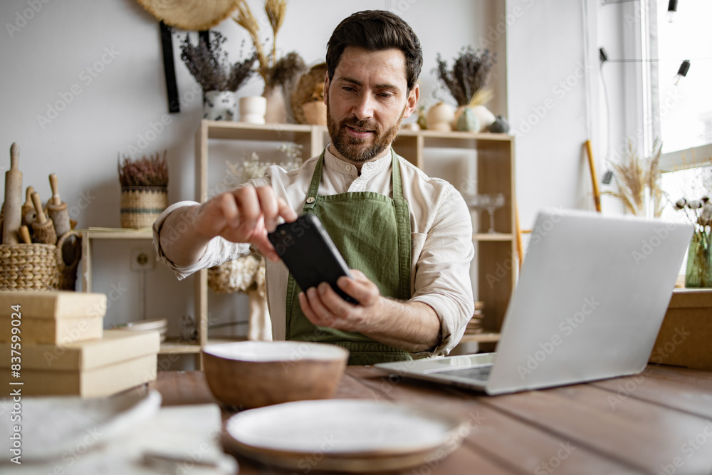 Mature man, potter, engaged in making home-made dishes and updating website portfolio. Positive manager of decorative clay store takes photo on phone for social media. Mature man, potter, engaged in making home-made dishes and updating website portfolio. Positive manager of decorative clay store takes photo on phone for social media.