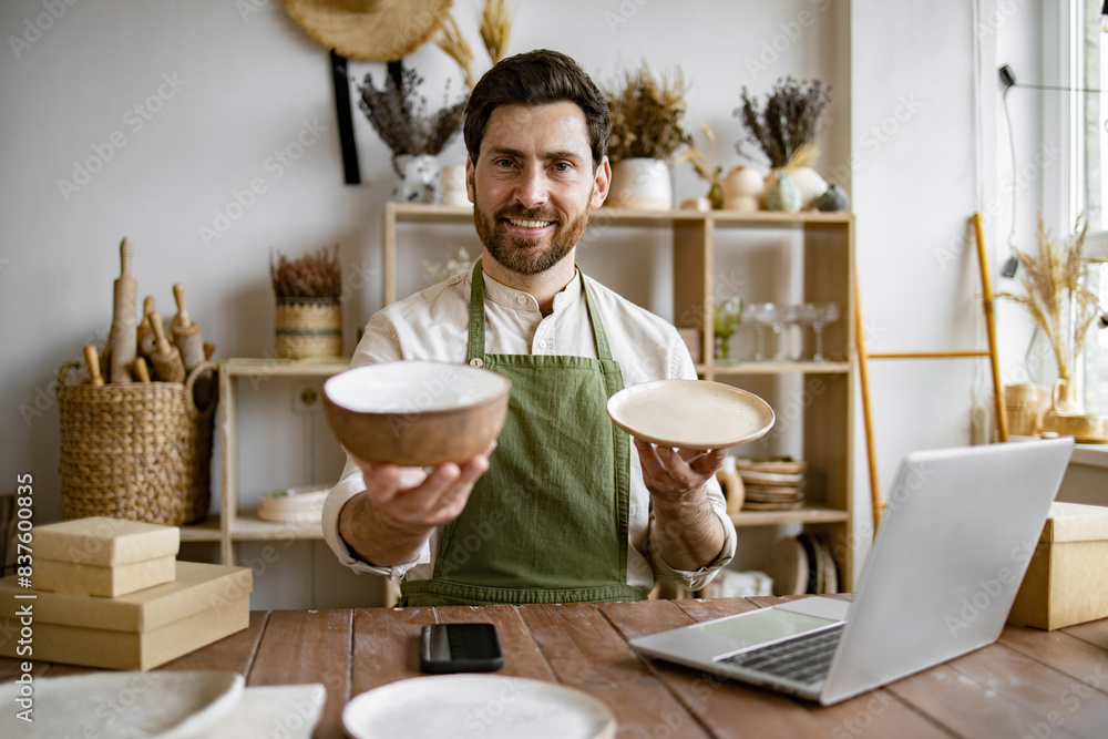 Attractive bearded man wearing green apron receiving goods at modern decor store. Professional seller checking for no damages on bowl and plate that holding in hands. Attractive bearded man wearing green apron receiving goods at modern decor store. Professional seller checking for no damages on bowl and plate that holding in hands.