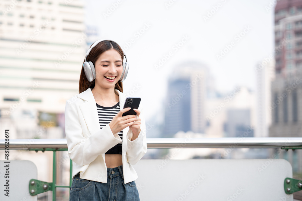 Young asian woman using smartphone while walking in the urban city. Happy Beautiful asian female with headphone listen to the music by cell phone standing at office building city. Lifestyle people Young asian woman using smartphone while walking in the urban city. Happy Beautiful asian female with headphone listen to the music by cell phone standing at office building city. Lifestyle people