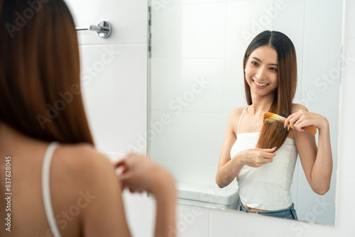 A beautiful woman with long hair is thinking about what style she should cut her hair with scissors or going to a beauty salon. Looking at mirror in restroom