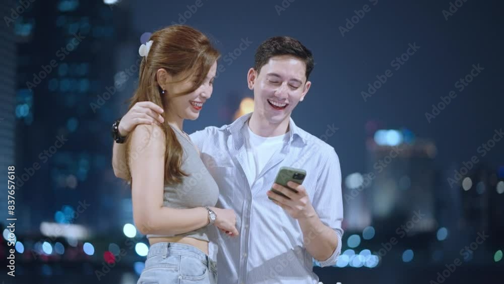 Attractive caucasian man flirting on date with happy smiling beautiful asian girlfriend, showing his mobile phone app through device screen while standing during late night in front of city skyscraper Attractive caucasian man flirting on date with happy smiling beautiful asian girlfriend, showing his mobile phone app through device screen while standing during late night in front of city skyscraper
