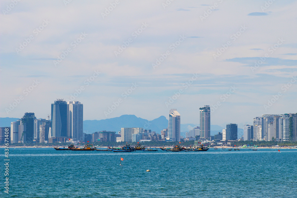 Photograph of Da Nang, Vietnam skyline viewed from the beach, with traditional fishing boats anchored near the shore Photograph of Da Nang, Vietnam skyline viewed from the beach, with traditional fishing boats anchored near the shore