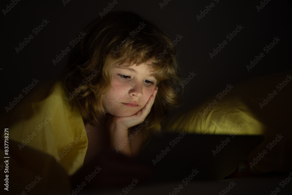 Kid is addicted to tablet, kid use tablet, watching cartoon. Child watching his tablet in the bed. Illuminated child face from device screen. Boy under the covers hold a tablet. Night time. Kid is addicted to tablet, kid use tablet, watching cartoon. Child watching his tablet in the bed. Illuminated child face from device screen. Boy under the covers hold a tablet. Night time.