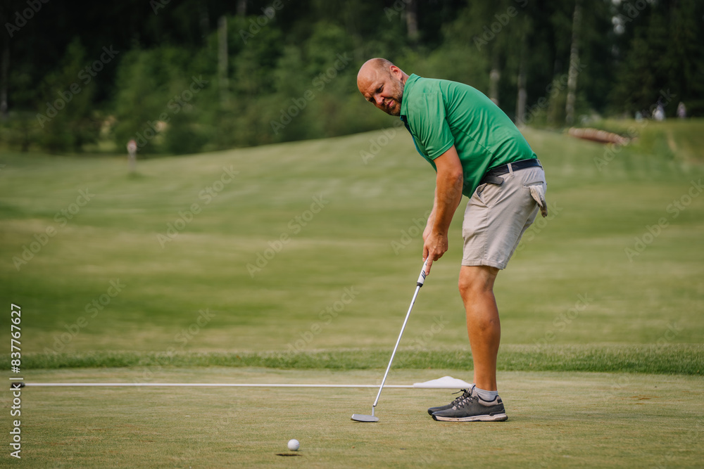 Fototapeta premium Valmiera, Latvia - June 1, 2024 - Man in a green shirt putting on a golf course on a sunny day, with trees and greenery in the background. Blurred background.