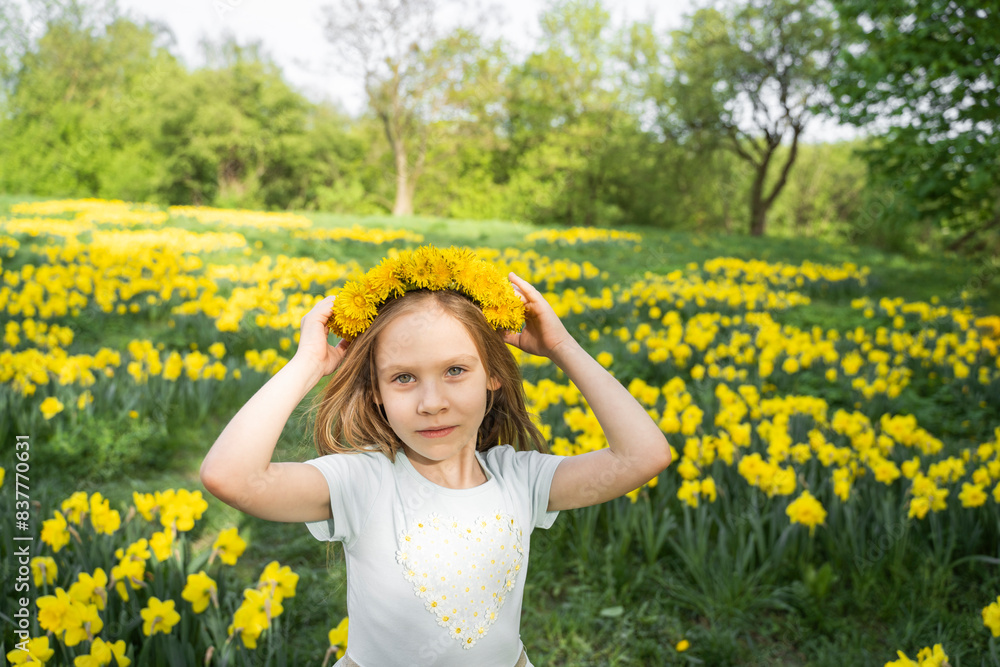 Smiling girl wearing dandelion wreath and standing near daffodil flowers in field Smiling girl wearing dandelion wreath and standing near daffodil flowers in field
