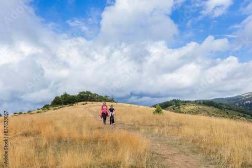 Spirit Of Adventure With This Scenic Image Of Mother And Daughter Hikers Walking Along A Dirt Path Surrounded By Lush Greenery And Framed By Distant Mountains