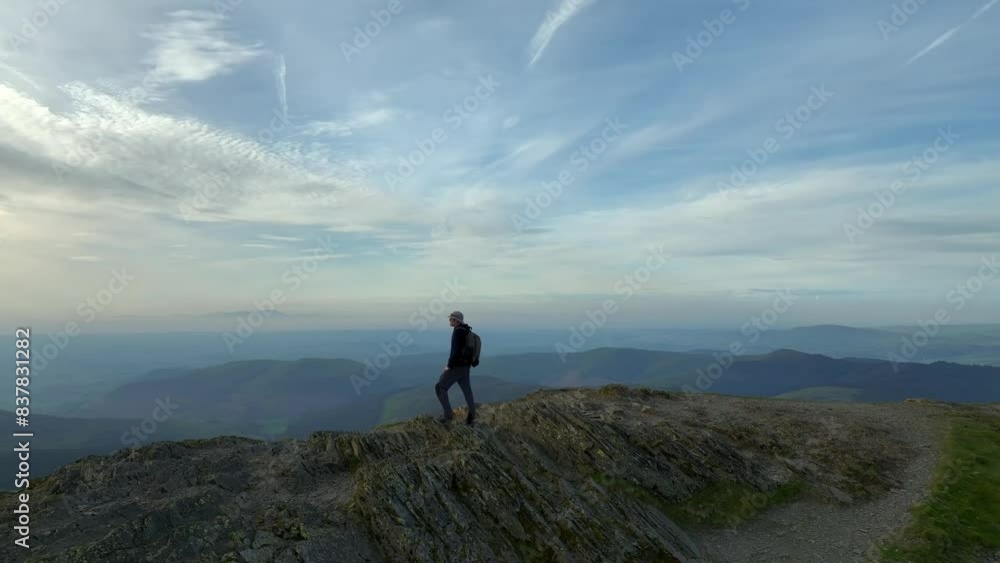 Mountain walker on craggy summit at golden hour. Camera 180 orbit revealing misty mountains and wide green valley with lake. Grisedale Pike, Lake District, Cumbria, England, UK Mountain walker on craggy summit at golden hour. Camera 180 orbit revealing misty mountains and wide green valley with lake. Grisedale Pike, Lake District, Cumbria, England, UK