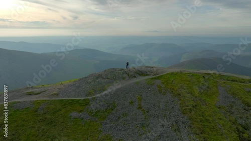 Solitary mountain walker on summit with wide slow orbit revealing misty valleys and mountains prior to sunset. Grisedale Pike, Lake District, Cumbria, England, UK photo