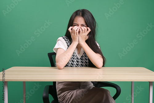 Young Woman Sitting On Desk And Covering Her Face With Hands Isolated Over Green Background