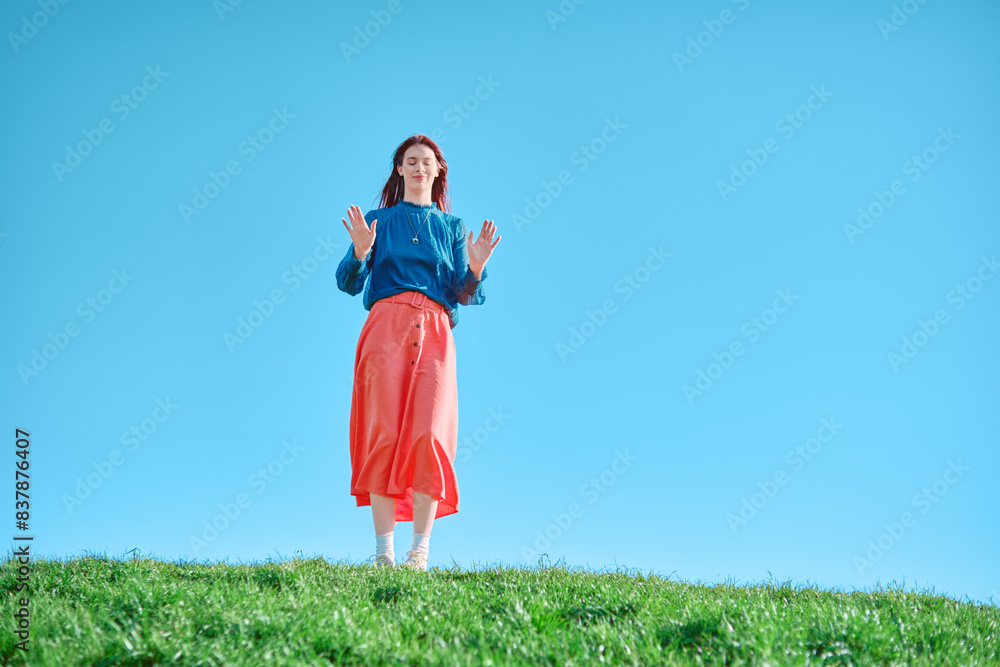 Teen girl in blue top and coral skirt standing on a grassy hill against a blue sky. Outdoor portrait with bright colors and clear sky. Low angle shot. Teen girl in blue top and coral skirt standing on a grassy hill against a blue sky. Outdoor portrait with bright colors and clear sky. Low angle shot.