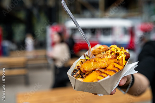 Close-up of hand holding street food paper tray with grilled items and toppings outdoors.