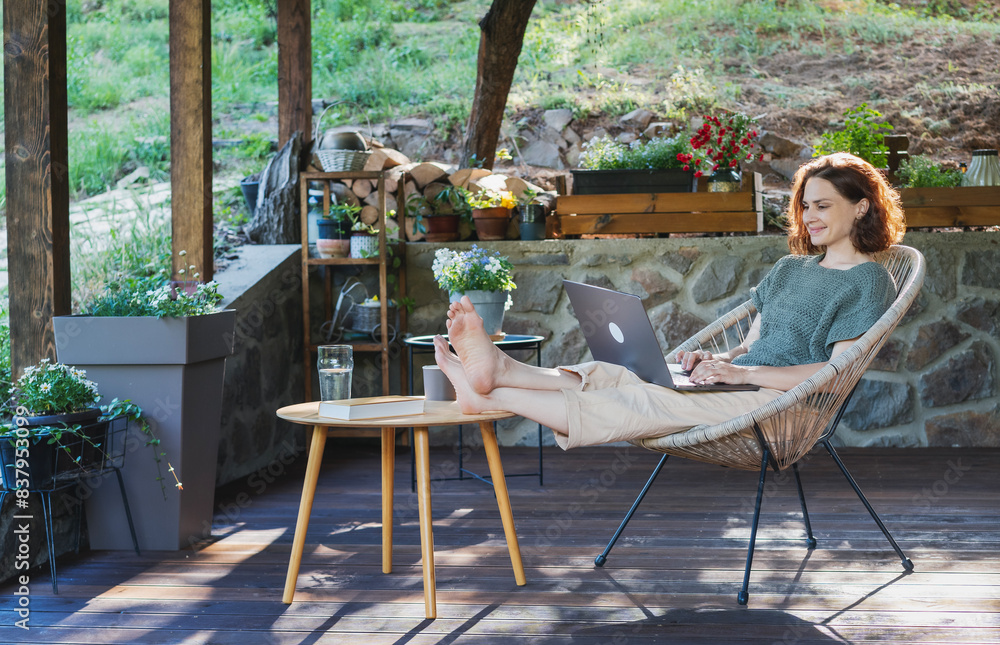 Young adult Caucasian woman sitting on cottage terrace using laptop in summertime, online work