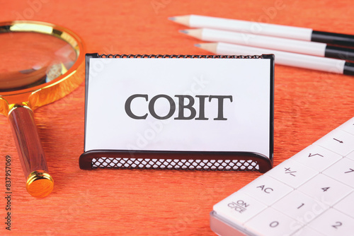 COBIT text on a white business card in a stand with a magnifying glass, pencils and a calculator photo