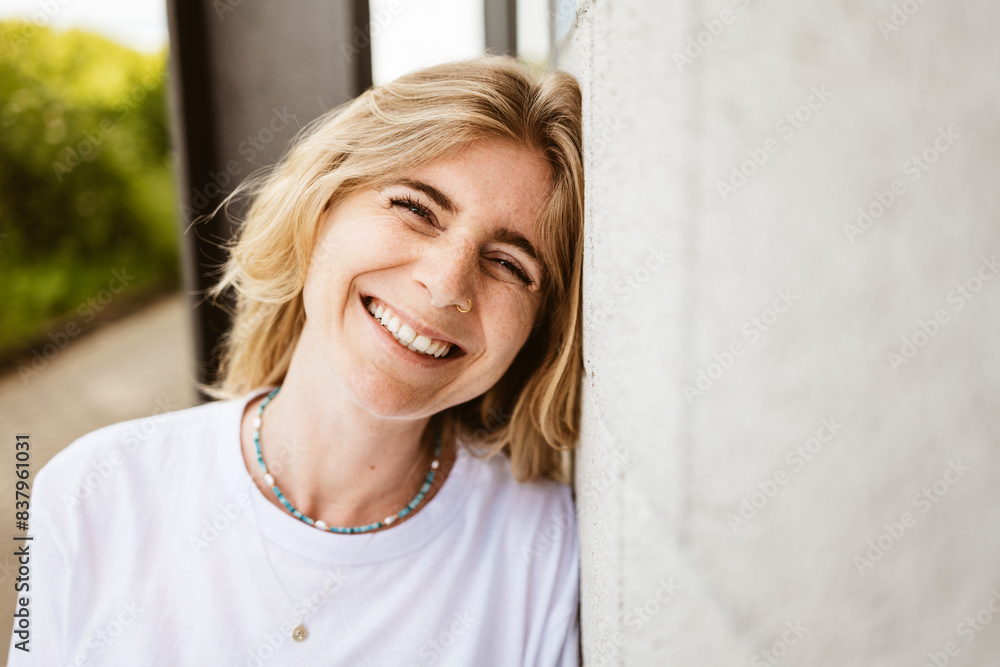Blonde woman with a bright smile, leaning against a wall outdoors near a modern building, exuding happiness and warmth.