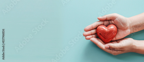 Adult and child hands holding a red heart on an aqua background symbolize heart health, donation, corporate social responsibility, World Heart Day, World Health Day, and Family Day.
