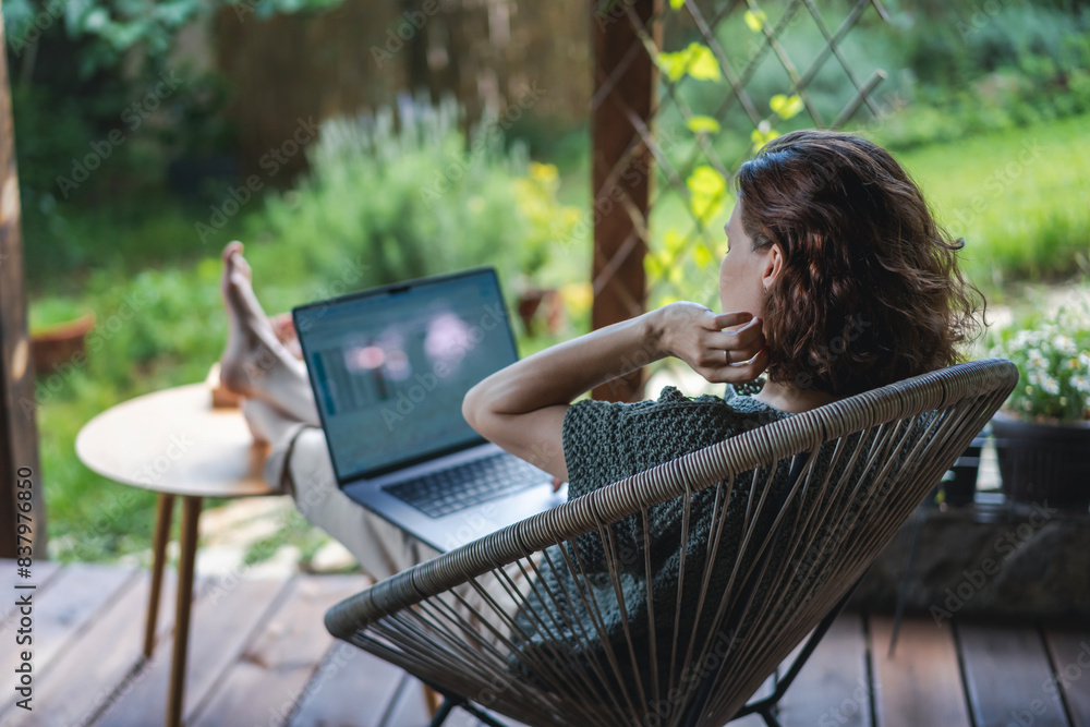 Young adult Caucasian woman sitting on cottage terrace using laptop in summertime, online work