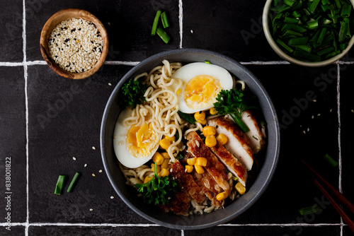 A delicious and hearty bowl of chicken ramen with a boiled egg, fresh green onion, and sweet corn, served over a bed of noodles in a flavorful broth. Black tile background