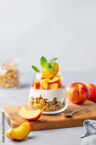 Greek yogurt parfait with peach and granola in a glass on a wooden board on a light background with fresh fruits.