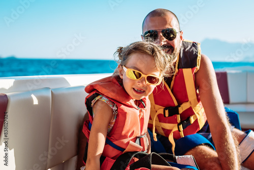 A little girl and her father, both wearing orange life jackets, seated on a boat, enjoying a recreational activity on the water as a family