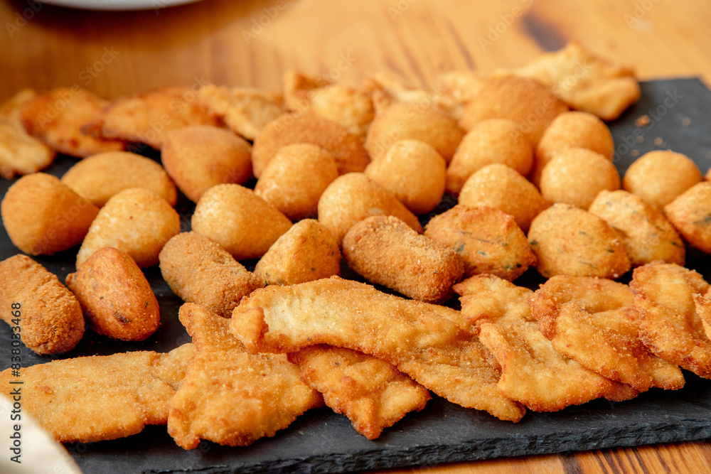 An inviting platter of assorted fried snacks, including golden-brown croquettes, chicken strips, and cheese balls, perfectly arranged on a slate board, ready to be enjoyed at a social gathering. An inviting platter of assorted fried snacks, including golden-brown croquettes, chicken strips, and cheese balls, perfectly arranged on a slate board, ready to be enjoyed at a social gathering.