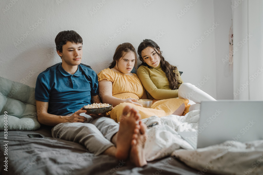 Down syndrome woman watching movie on laptop while sitting with siblings on bed at home Down syndrome woman watching movie on laptop while sitting with siblings on bed at home